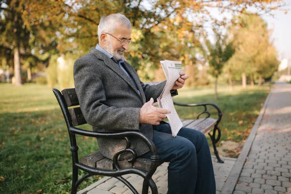 Hombre mayor barbudo en gafas leyendo periódico en el parque de otoño. Hombre guapo de pelo gris sentado en el banco temprano en la mañana —  Fotos de Stock