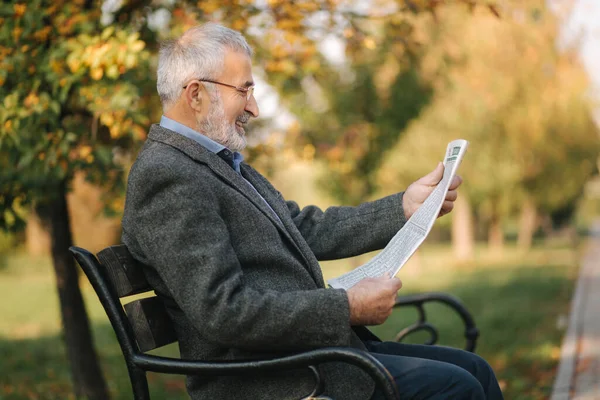 Homme âgé barbu dans des lunettes de lecture journal dans le parc d'automne. Beau homme aux cheveux gris assis sur le banc tôt le matin — Photo