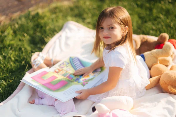 Очаровательная маленькая девочка сидит на клетчатой улице и читает книгу. Счастливая девочка проводит время на заднем дворе на закате — стоковое фото