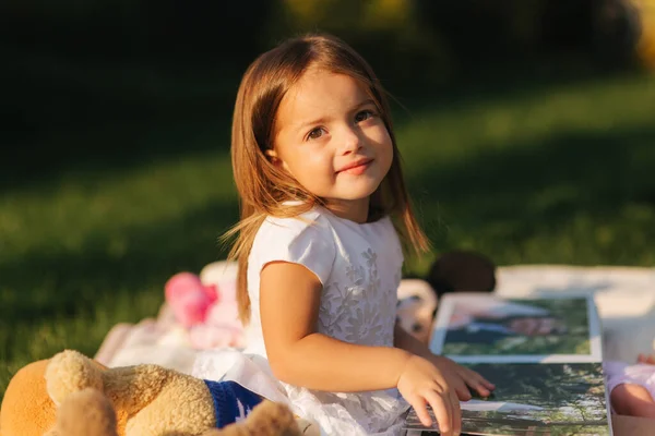 Αξιολάτρευτο κοριτσάκι κάθεται σε καρό ύπαιθρο και να διαβάσετε το βιβλίο. Ευτυχισμένο χαμογέλασε κοριτσάκι περνούν το χρόνο τους στην πίσω αυλή στο ηλιοβασίλεμα — Φωτογραφία Αρχείου
