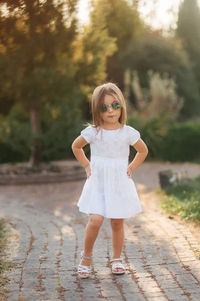 Милая маленькая девочка в белом платье позирует фотографу. Счастливый малыш в солнечных очках — стоковое фото