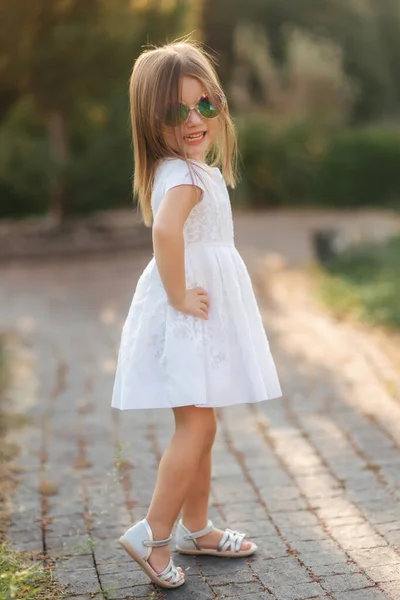 Χαριτωμένο κοριτσάκι με λευκό φόρεμα που ποζάρει στον φωτογράφο. Ευτυχισμένο μικρό παιδί με γυαλιά ηλίου — Φωτογραφία Αρχείου