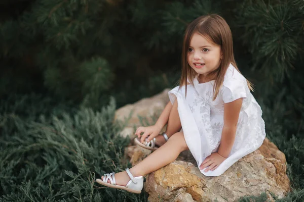 Klein meisje in witte jurk zit op grote steen in de voorkant van groene naaldboom — Stockfoto
