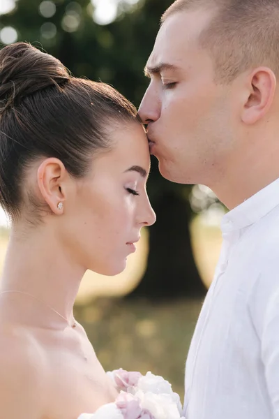 Ritratto ravvicinato di sposo baciare la sposa in fronte davanti a un grande albero. Vista laterale — Foto Stock