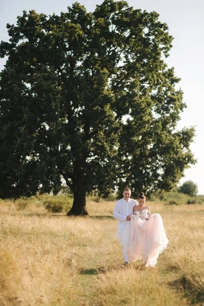 Felizes recém-casados a entrar junto à grande árvore no campo. — Fotografia de Stock