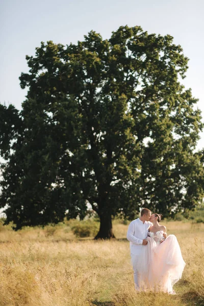 Boldog friss házasok sétálnak be a nagy fa mellett a mezőn. — Stock Fotó