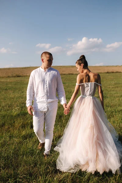 Gorgeous bride with handsome groom walkin in the field after wedding ceremony. Newlyweds posing to photographer — Stock Photo, Image