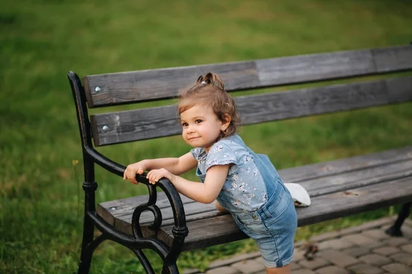 Cute little girl w dżinsach wspina się na ławce w parku. Szczęśliwy uśmiechnięty dzieciak na ławce. — Zdjęcie stockowe