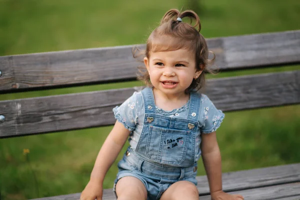 Schattig klein meisje dat lacht. Mooi vrouwelijk kind in denim zittend op de bank in het park. Gelukkige kleine meid glimlach. 18 maanden — Stockfoto