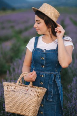 Yaz lavanta tarlasında yürüyen çekici genç bir kadın. Hasır şapka ve çantalı kot elbise giymiş bir manken.