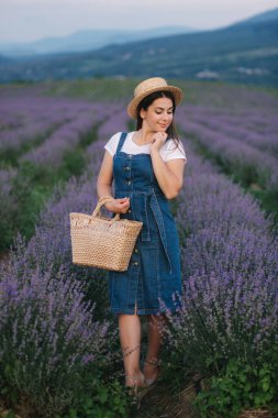 Yaz lavanta tarlasında yürüyen çekici genç bir kadın. Hasır şapka ve çantalı kot elbise giymiş bir manken.