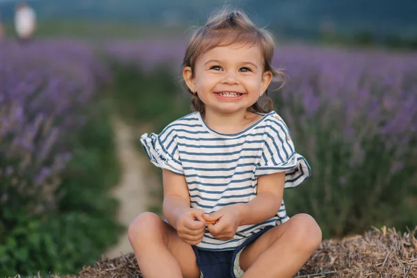 사랑스럽게 웃는 어린 소녀의 모습이 농장 옆 건초 위에 앉아 있다. 여름 라벤더들 판의 배경. 줄무늬 티셔츠와 파란 반바지를 입은 귀여운 소녀 — 스톡 사진