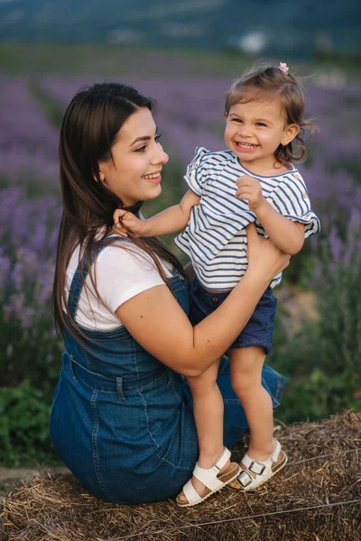 Mamma e figlioletta si siedono sul fieno vicino alla fattoria. Sfondo del campo estivo di lavanda. Stile denim di famiglia. Bambina abbraccio mamma — Foto Stock