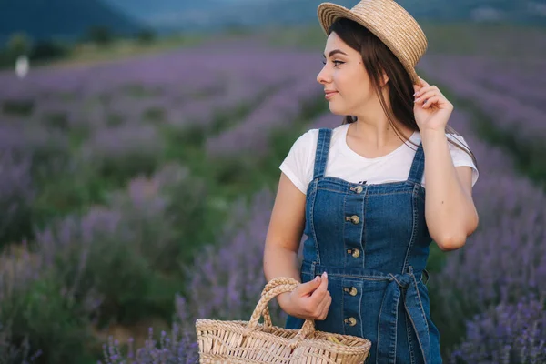 Atractiva joven mujer caminando en el campo de lavanda de verano. Modelo vestido con vestido de mezclilla con sombrero de paja y bolsa — Foto de Stock
