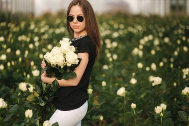 Elinde buket beyaz gül olan genç bir kadın seradaki gül tarlasının içinde duruyor.