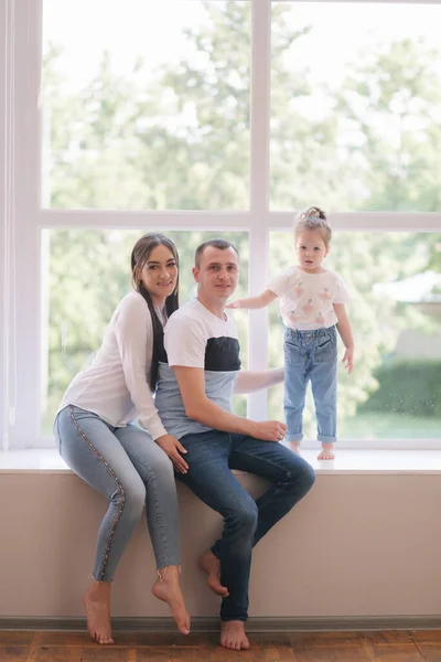 Gadis kecil dengan ibu dan ayah duduk dekat jendela besar di studio. Foto keluarga — Stok Foto