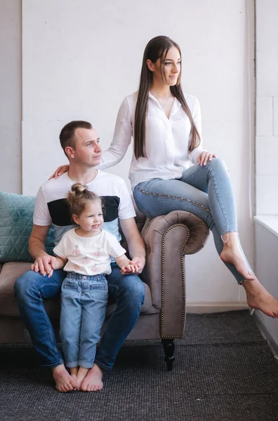 Fotosessão familiar em estúdio. Pequena filha mãe e pai se senta no sofá pela janela — Fotografia de Stock