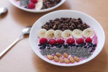 Taze ahududu, yabanmersini, chia tohumu, badem, muz ve çikolata parçacıklı meyve suyu kasesi. Sabah kahvaltısı ahşap masada. İki masal Smoothie reverans yapar