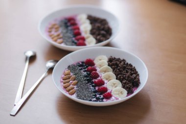 Taze ahududu, yabanmersini, chia tohumu, badem, muz ve çikolata parçacıklı meyve suyu kasesi. Sabah kahvaltısı ahşap masada. İki masal Smoothie reverans yapar