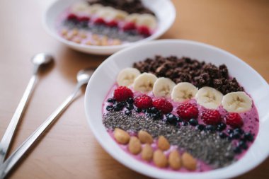 Taze ahududu, yabanmersini, chia tohumu, badem, muz ve çikolata parçacıklı meyve suyu kasesi. Sabah kahvaltısı ahşap masada. İki Ptales Fith Smoothie yayları. Vejetaryen yemeği