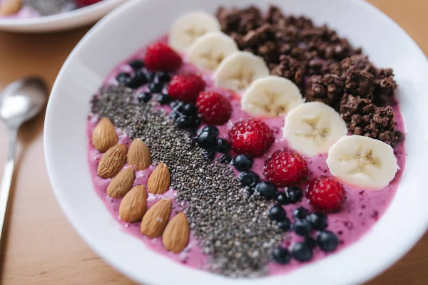 Smoothie bowl with fresh raspberry, blueberry, chia seeds, almonds, banana, and chocolate flakes. Morning breakfast on wooden table. Close up — Stock Photo, Image