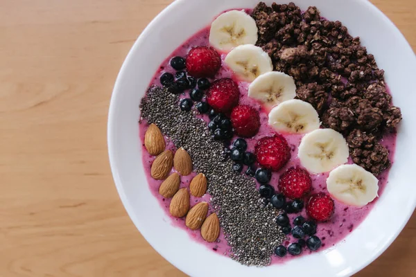 Smoothie bowl with fresh raspberry, blueberry, chia seeds, almonds, banana, and chocolate flakes. Morning breakfast on wooden table. Top view — Stock Photo, Image
