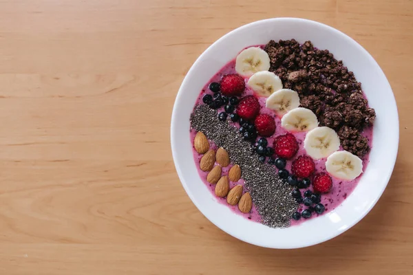 Smoothie bowl with fresh raspberry, blueberry, chia seeds, almonds, banana, and chocolate flakes. Morning breakfast on wooden table. Top view — Stock Photo, Image