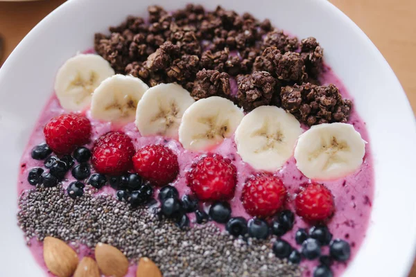 Smoothie bowl with fresh raspberry, blueberry, chia seeds, almonds, banana, and chocolate flakes. Morning breakfast on wooden table. Top view — Stock Photo, Image