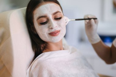 Kozmetik uzmanının elleri beyaz yüz maskesi takıyor. Deriyi sulandırır, yüzü parlar ve cildi parlatır. Çamur maskeli Hulf suratı. Çekici genç esmer, gözleri kapalı rahatlıyor.