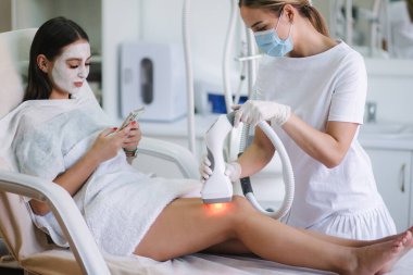 Beyaz maskeli güzel bir kadın telefonunu kullanırken beyaz eldivenli bir kozmetik uzmanı midesine radyasyon kaldırma prosedürü uyguluyor. Klinikteki güzel kadın. Güzellik uzmanı