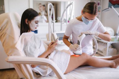 Beyaz maskeli güzel bir kadın telefonunu kullanırken beyaz eldivenli bir kozmetik uzmanı midesine radyasyon kaldırma prosedürü uyguluyor. Klinikteki güzel kadın. Güzellik uzmanı