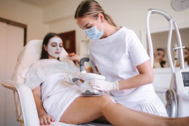 Çekici kadın dinlenirken beyaz eldivenli kozmetik uzmanı midesine radyasyon kaldırma prosedürü uyguluyor. Klinikteki güzel kadın. Tıbbi maskeli güzellik uzmanı.