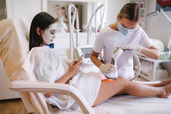 Beyaz maskeli güzel bir kadın telefonunu kullanırken beyaz eldivenli bir kozmetik uzmanı midesine radyasyon kaldırma prosedürü uyguluyor. Klinikteki güzel kadın. Güzellik uzmanı