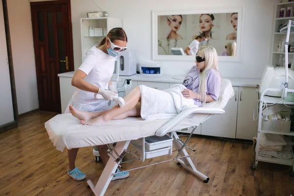 Beautician in medical mask and goggles doing epilation on beautiful girls leg in medical center. Female receiving laser light hair removal treatment for hairless smooth skin at cosmetology salon