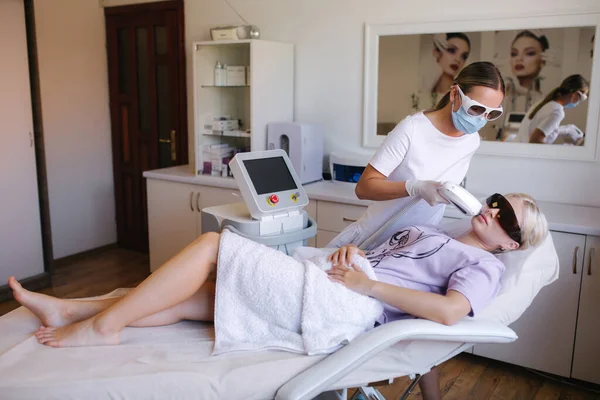 Beautician in medical mask doing epilation on beautiful womans face in medical center. Female receiving laser light hair removal treatment for hairless smooth skin at cosmetology salon