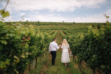 Damat ve gelin düğün günlerinde üzüm bağında yürüyorlar. Beyaz elbiseli sarışın kadın elinde düğün buketi tutuyor.