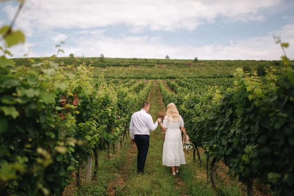 Bräutigam und Braut bei ihrem Hochzeitstag im Weinberg. Blonde Frau im weißen Kleid hält Brautstrauß in den Händen — Stockfoto