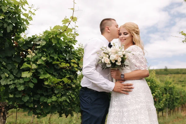 Bruidegom en bruid wandelen in de wijngaard op hun trouwdag. Blond haar vrouw in witte jurk houden bruiloft boeket in handen — Stockfoto