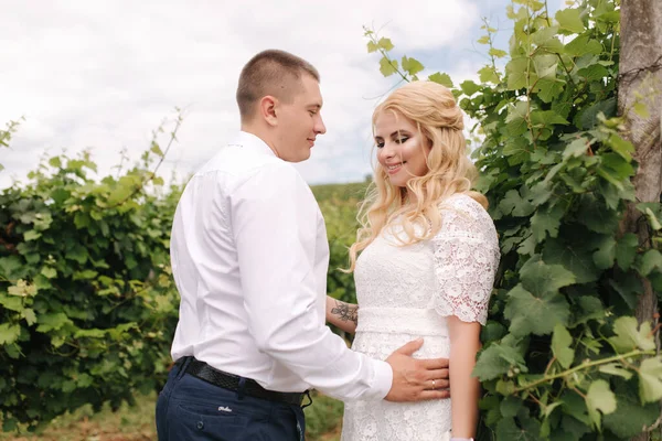 Bruidegom en bruid wandelen in de wijngaard op hun trouwdag. Blond haar vrouw in witte jurk houden bruiloft boeket in handen — Stockfoto