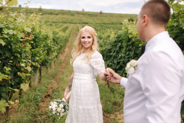 Brudgummen och bruden går i vingård på sin bröllopsdag. Blond hår kvinna i vit klänning hålla bröllop bukett i händerna — Stockfoto
