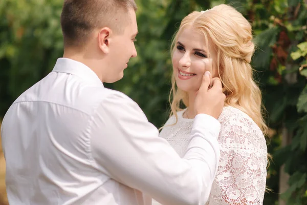 Brudgummen och bruden går i vingård på sin bröllopsdag. Blond hår kvinna i vit klänning hålla bröllop bukett i händerna — Stockfoto