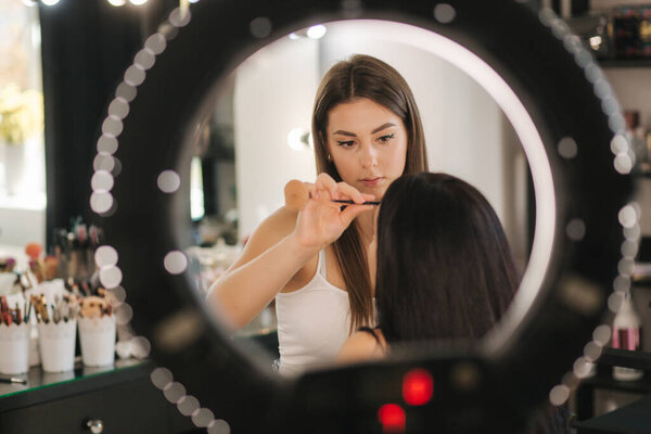 Photo though the ring lamp of makeup artist making a makeup for attractive young girl. Beautiful young brunette woman