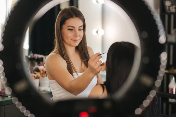 Makyaj sanatçısının yüzük lambası çekici genç bir kız için makyaj yapıyor. Güzel, genç esmer kadın. — Stok fotoğraf