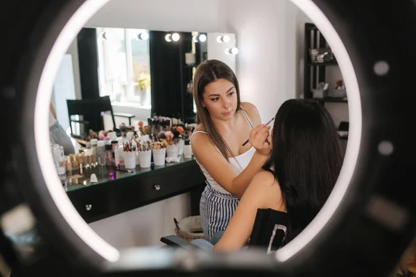 Makyaj sanatçısının yüzük lambası çekici genç bir kız için makyaj yapıyor. Güzel, genç esmer kadın. — Stok fotoğraf