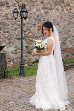 Zarif gelin, buketi taş duvarın önünde elinde tut.