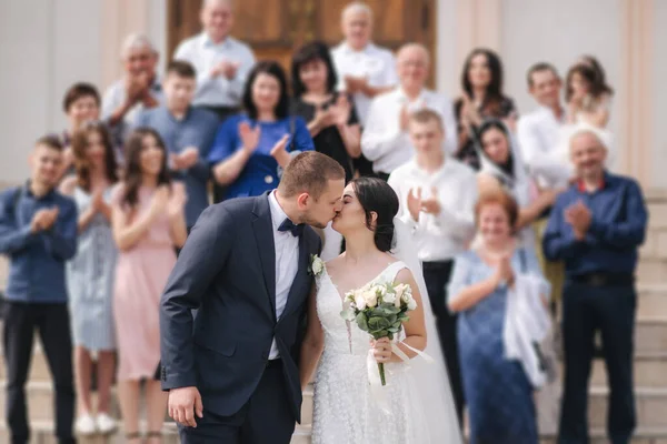 Noivo e noiva após a cerimônia de casamento pela igreja beijar uns aos outros na frente da família — Fotografia de Stock