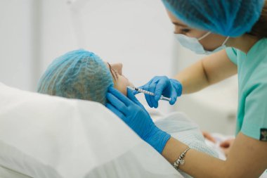 Kozmetik uzmanının elleri yüzüne ve dudaklarına iğne yapıyor. Genç bir kadın, güzellik kliniğinde yüz aşısı yaptırıyor. Yaşlanma, gençleşme ve hidrasyon prosedürleriyle yüzleş. Estetik kozmetoloji. Güzellik salonu