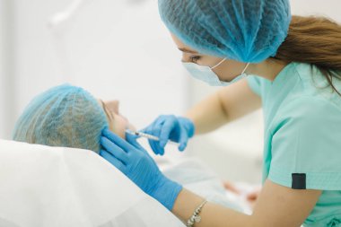 Kozmetik uzmanının elleri yüzüne ve dudaklarına iğne yapıyor. Genç bir kadın, güzellik kliniğinde yüz aşısı yaptırıyor. Yaşlanma, gençleşme ve hidrasyon prosedürleriyle yüzleş. Estetik kozmetoloji. Güzellik salonu