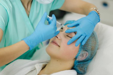 Kozmetik uzmanının elleri yüzüne ve dudaklarına iğne yapıyor. Genç bir kadın, güzellik kliniğinde yüz aşısı yaptırıyor. Yaşlanma, gençleşme ve hidrasyon prosedürleriyle yüzleş. Estetik kozmetoloji. Güzellik salonu