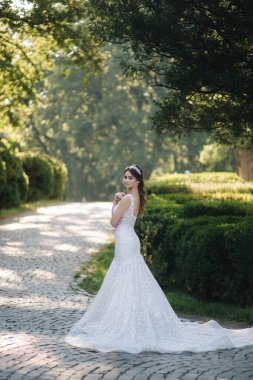 Lüks gelinlik içinde, büyük trenli, açık havada bekleyen muhteşem bir gelin. Yeşil ağacın arkaplanı